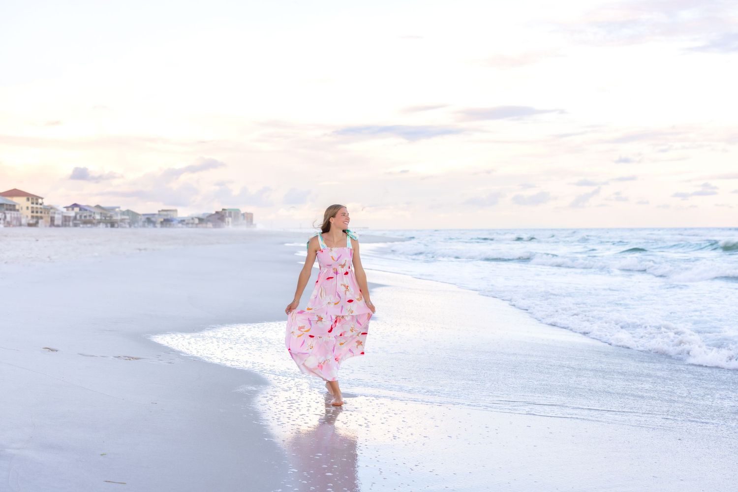 A person in a flowing pink dress walks along a white sandy beach at sunset with waves rolling in.