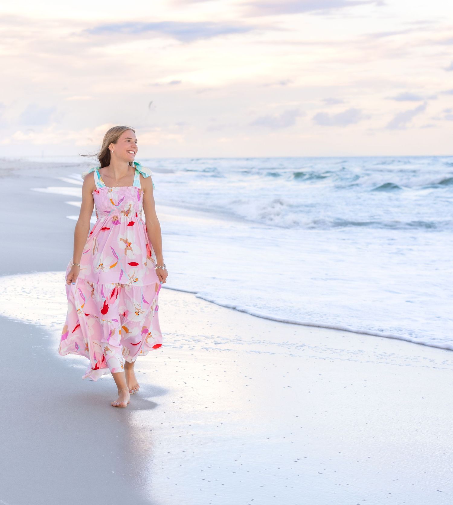Person in a floral print dress walks along a white sandy beach with ocean waves in the background at sunset.