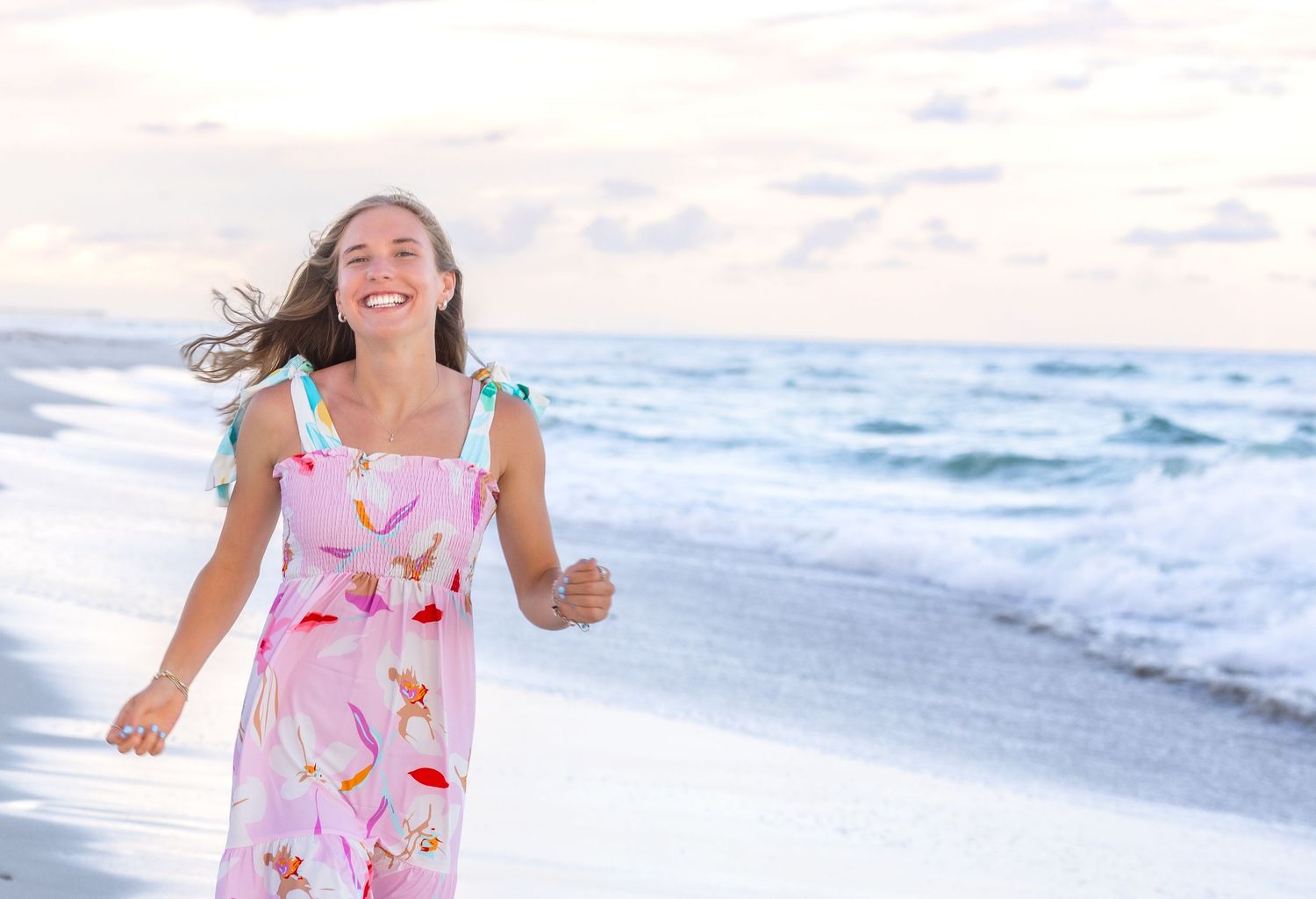 Joyful person in floral summer dress running along beach with waves and cloudy sky in background.