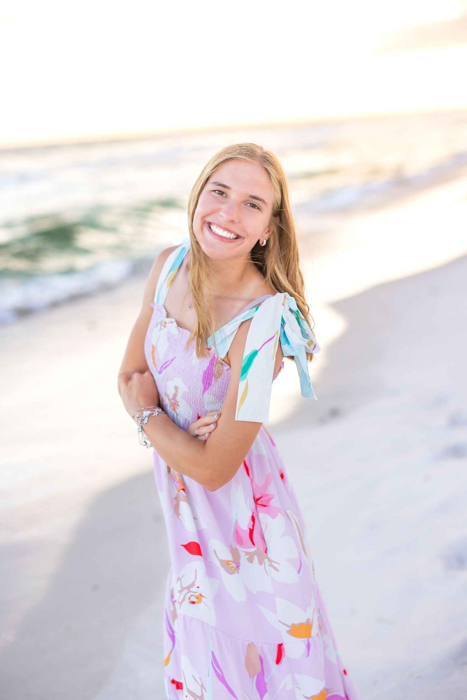 A person in a pink floral dress smiles while standing on a beach at sunset.