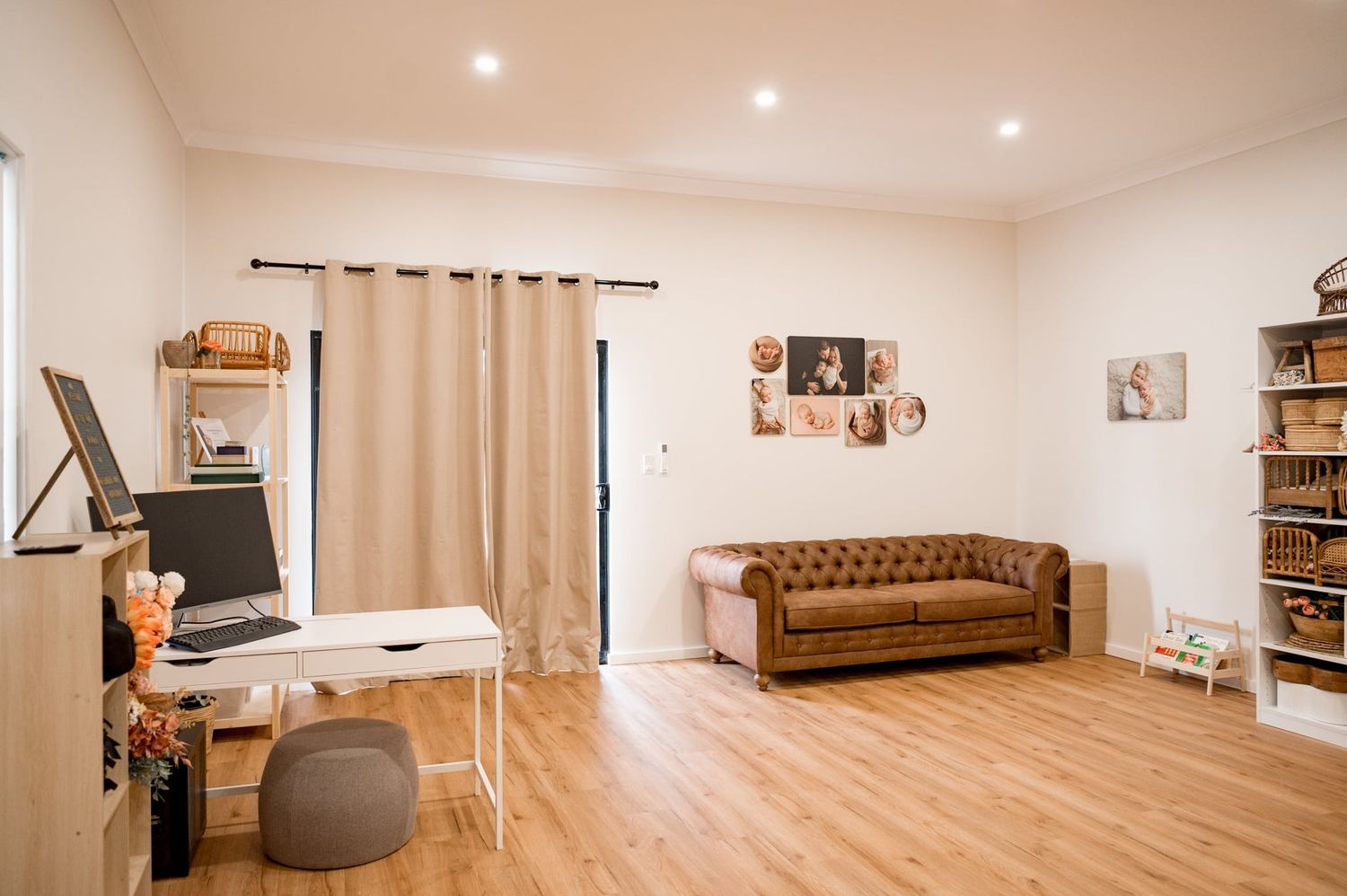 A modern home office space with a brown leather Chesterfield sofa, white desk, and wooden floating shelves on neutral walls.