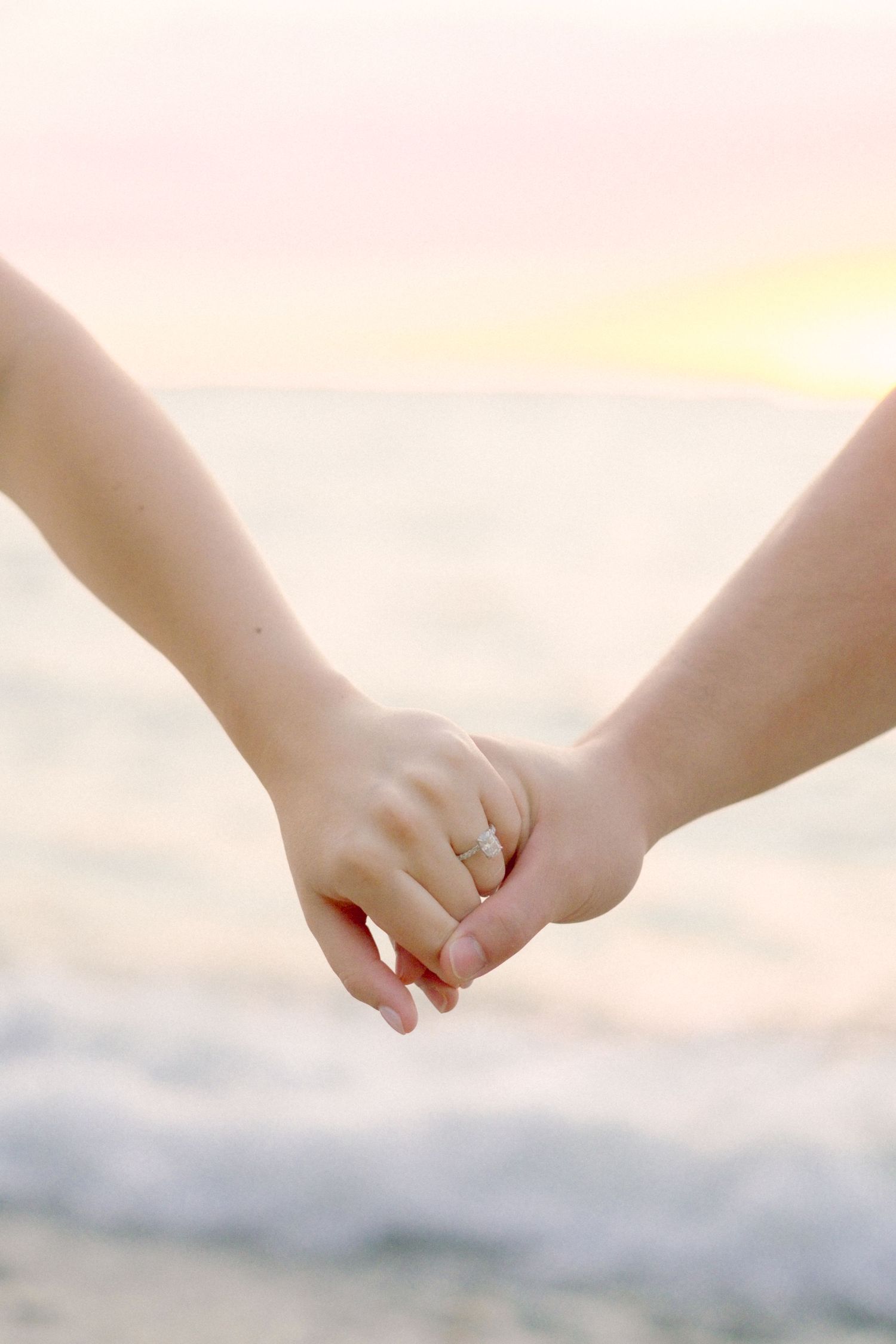Delicate intertwined hands photographed against a soft sunset backdrop.