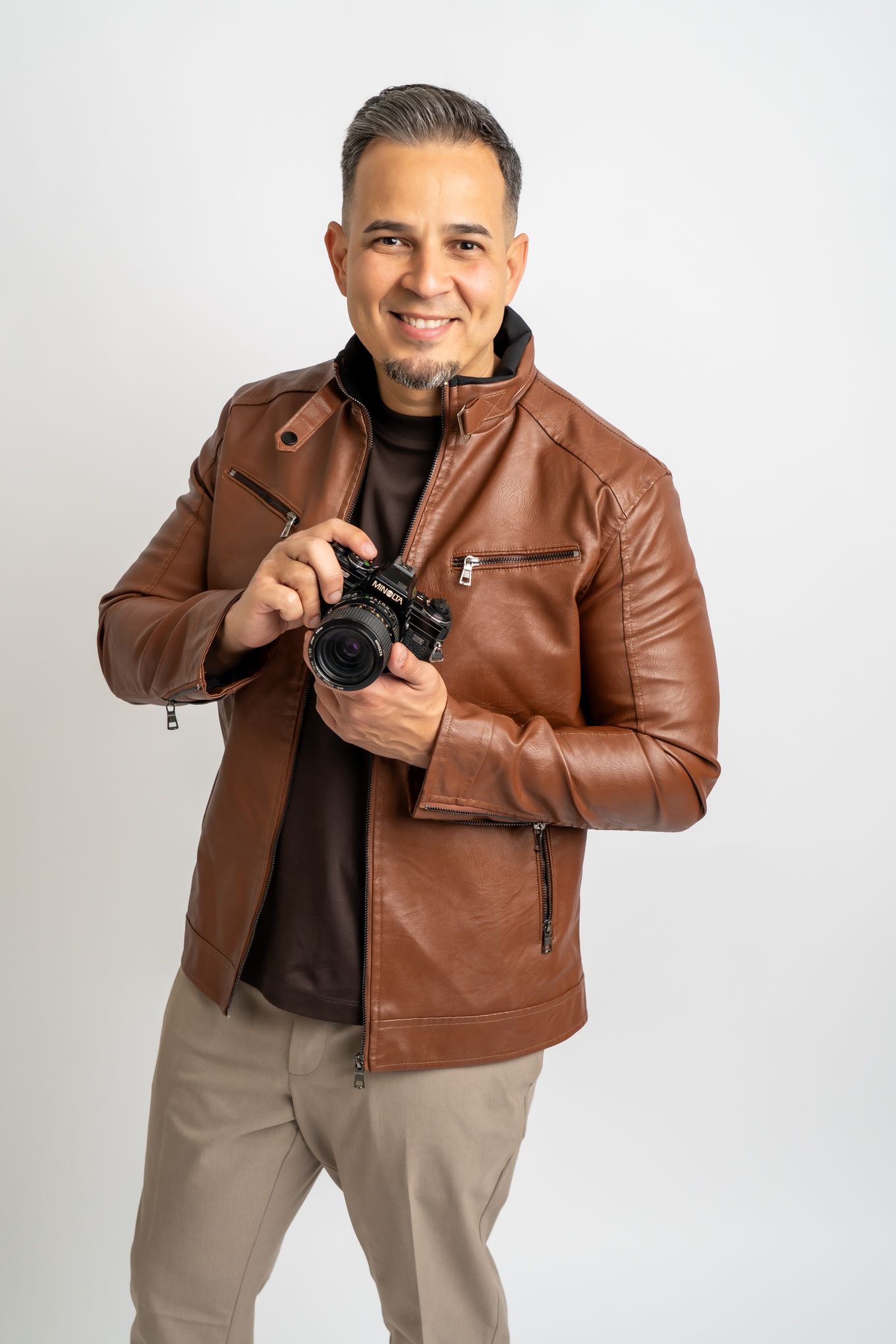 Photographer in brown leather jacket and khaki pants holding a digital camera against white background.
