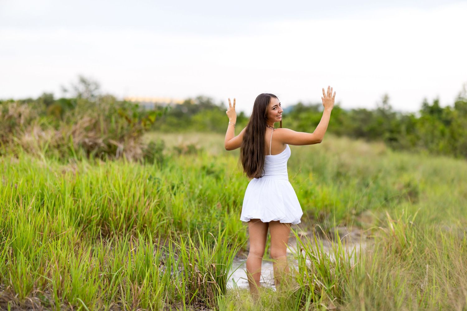 Woman in white dress poses with raised arms in tall grass field during golden hour.