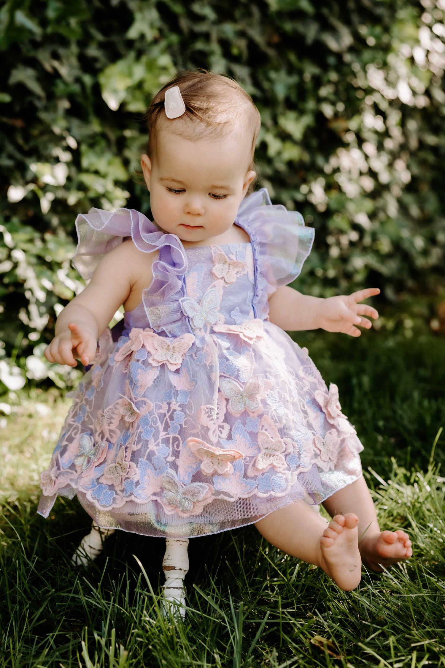 Baby wearing delicate purple and pink floral dress sits on grass in garden setting.