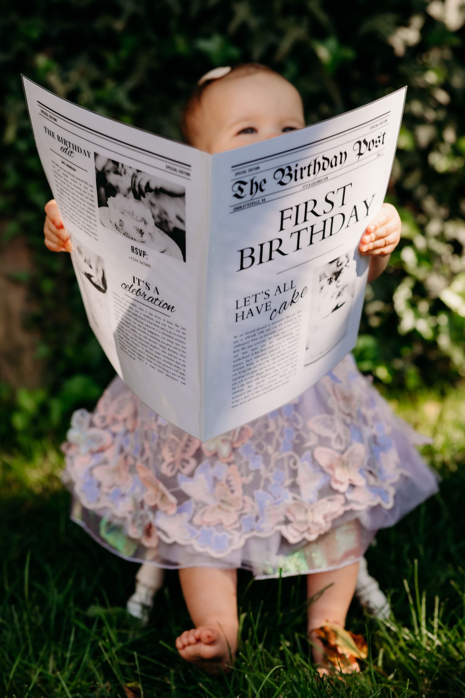 A small child in a tulle dress sits on grass while holding a custom newspaper titled 'The Birthday Post' for their first birthday.