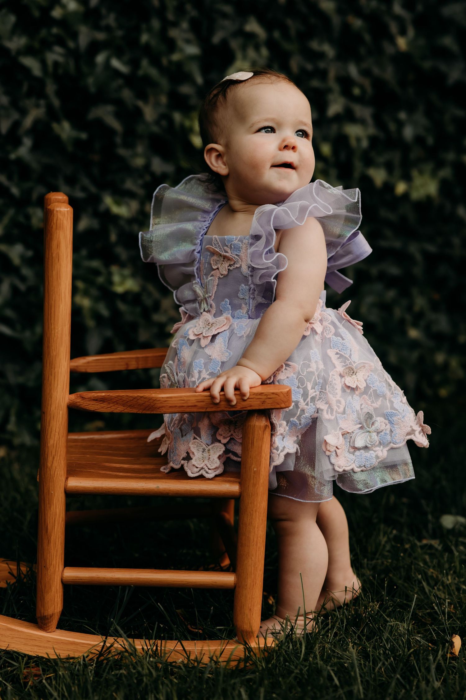 A baby in a frilly lavender dress stands beside a wooden rocking chair in a garden setting.