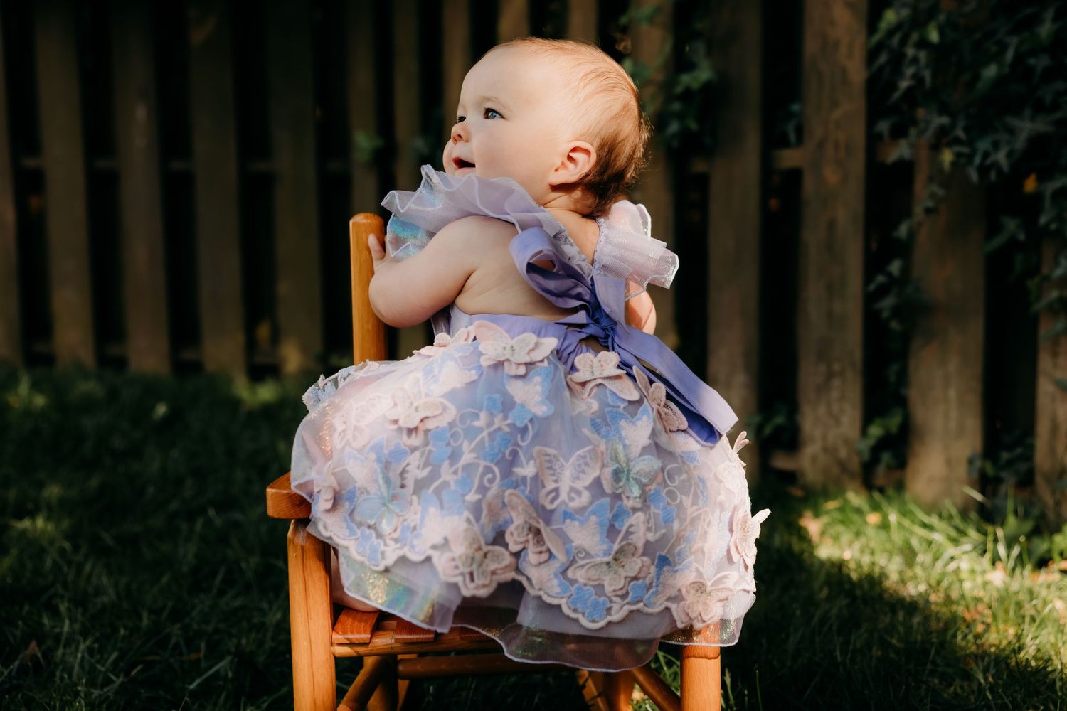 Small child in a frilly lavender dress sits on a wooden chair in a backyard setting during golden hour.