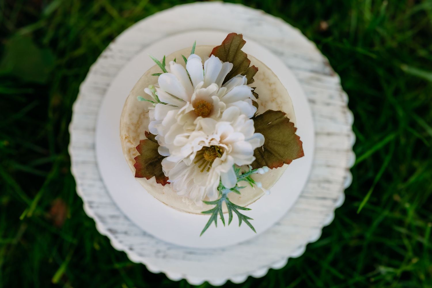 White daisies and greenery arranged on a small decorative cake with scalloped white stand placed on grass.