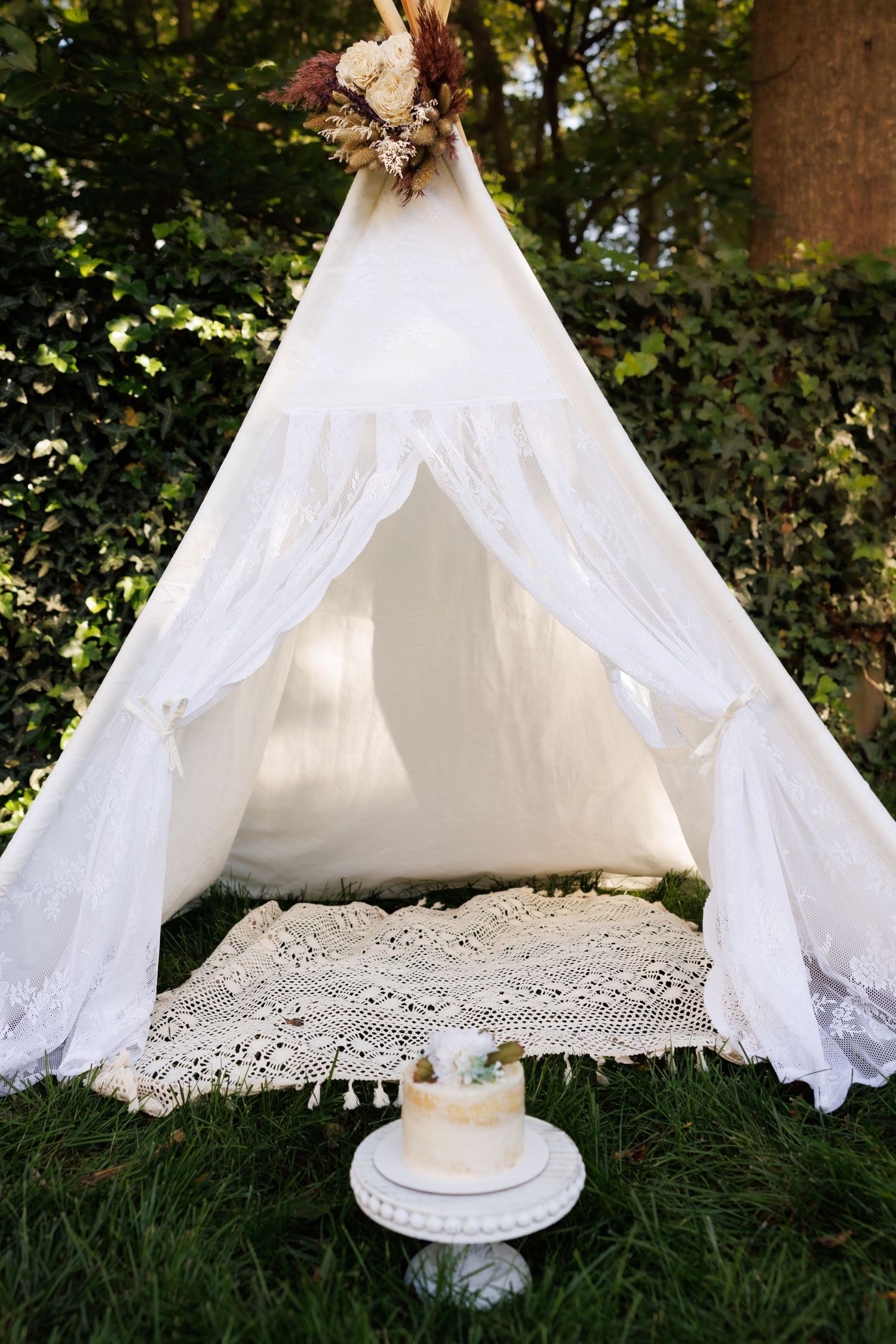 A romantic white lace teepee tent setup with a crochet rug and decorative accents in an outdoor garden setting.