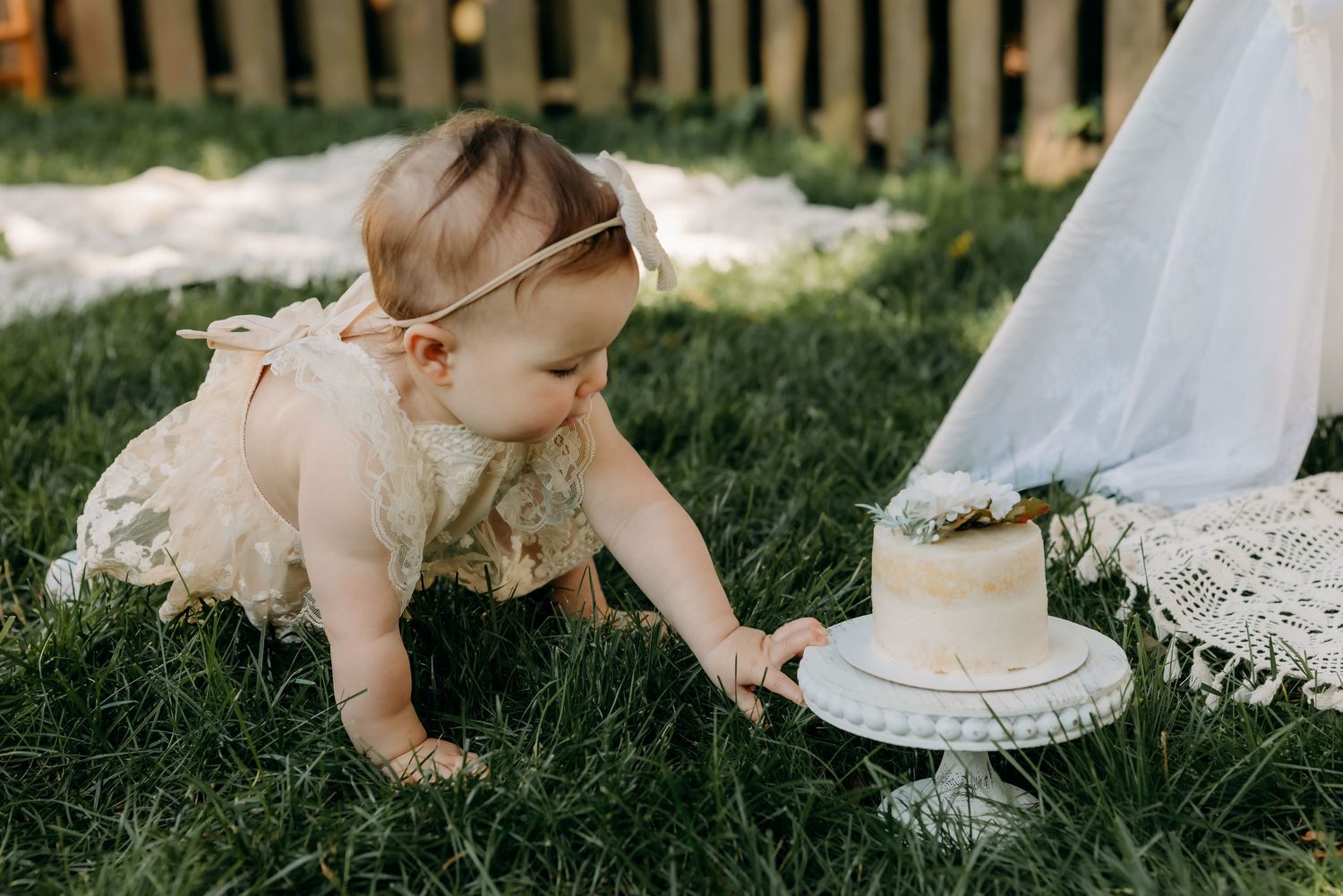 Small child in lace dress reaching for white cake on pedestal stand during outdoor cake smash session on grass.