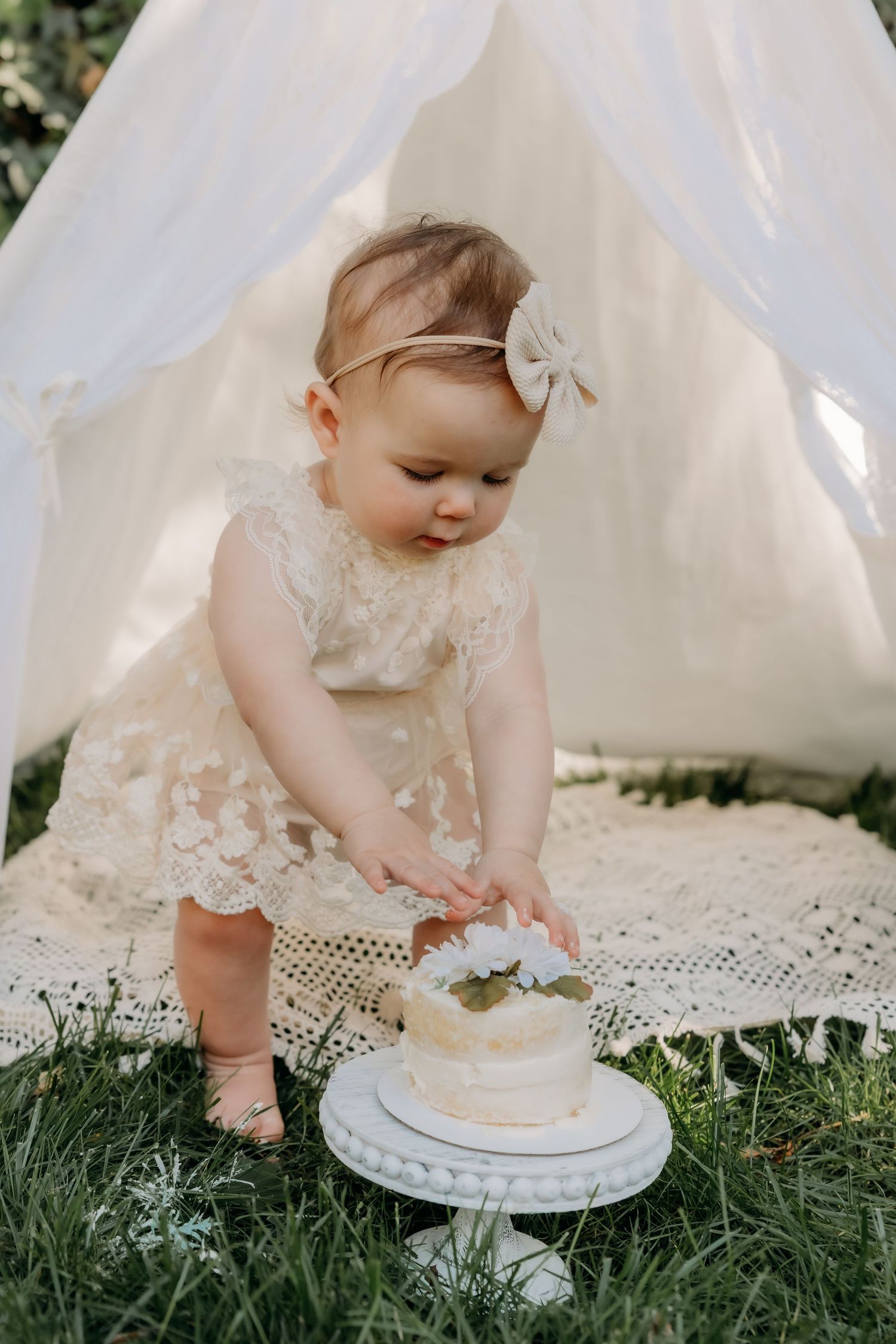 Baby in lace dress reaches for small white cake while sitting on lace blanket under white tent canopy outdoors.