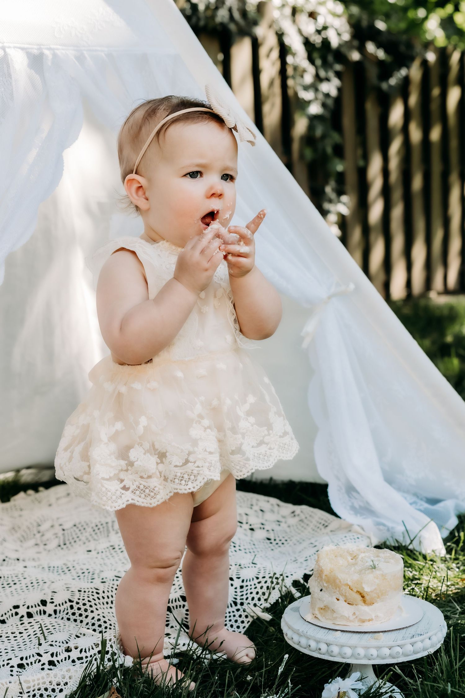 A baby wearing a lace dress stands next to a teepee and cake during an outdoor photoshoot in a garden setting.