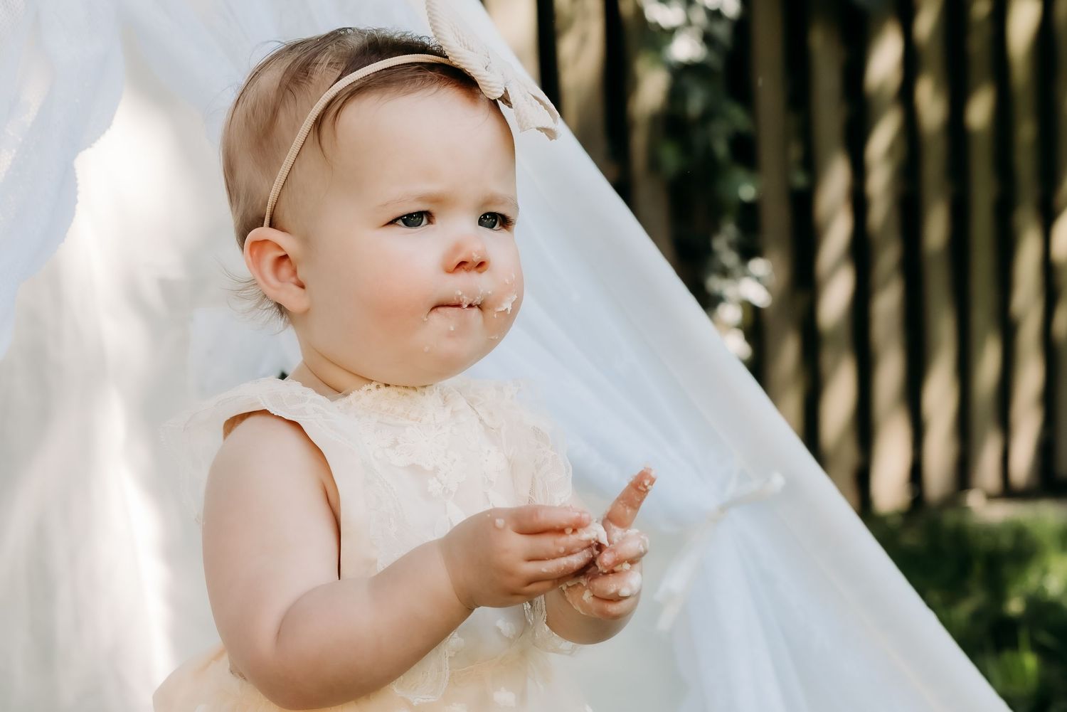 Baby wearing a white sleeveless dress stands next to white fabric outdoors on a sunny day.