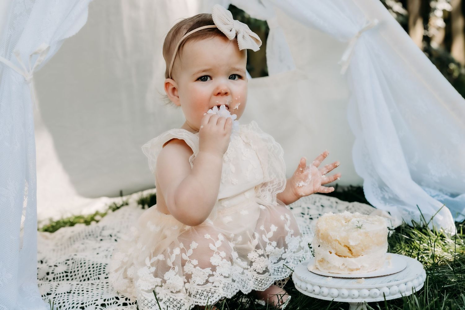 Baby in white lace dress sits on blanket outdoors enjoying birthday cake smash photo session.