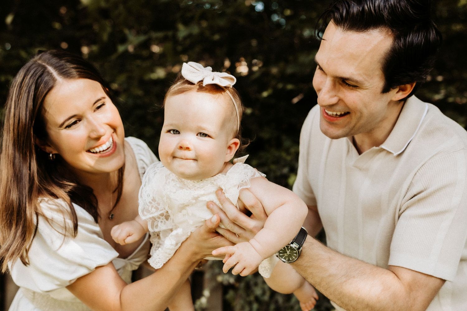 A family shares a joyful moment outdoors together with their baby wearing a white dress and bow headband.