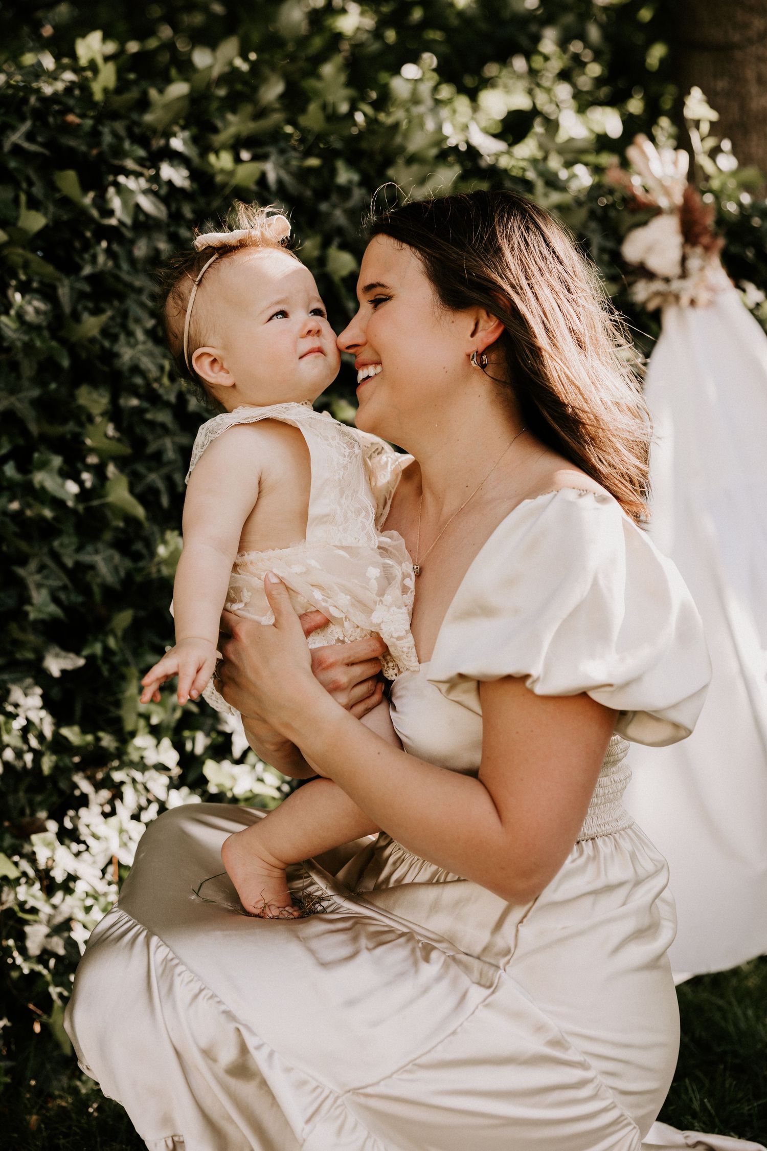 A tender moment between parent and baby in white dresses against a leafy backdrop in warm sunlight.