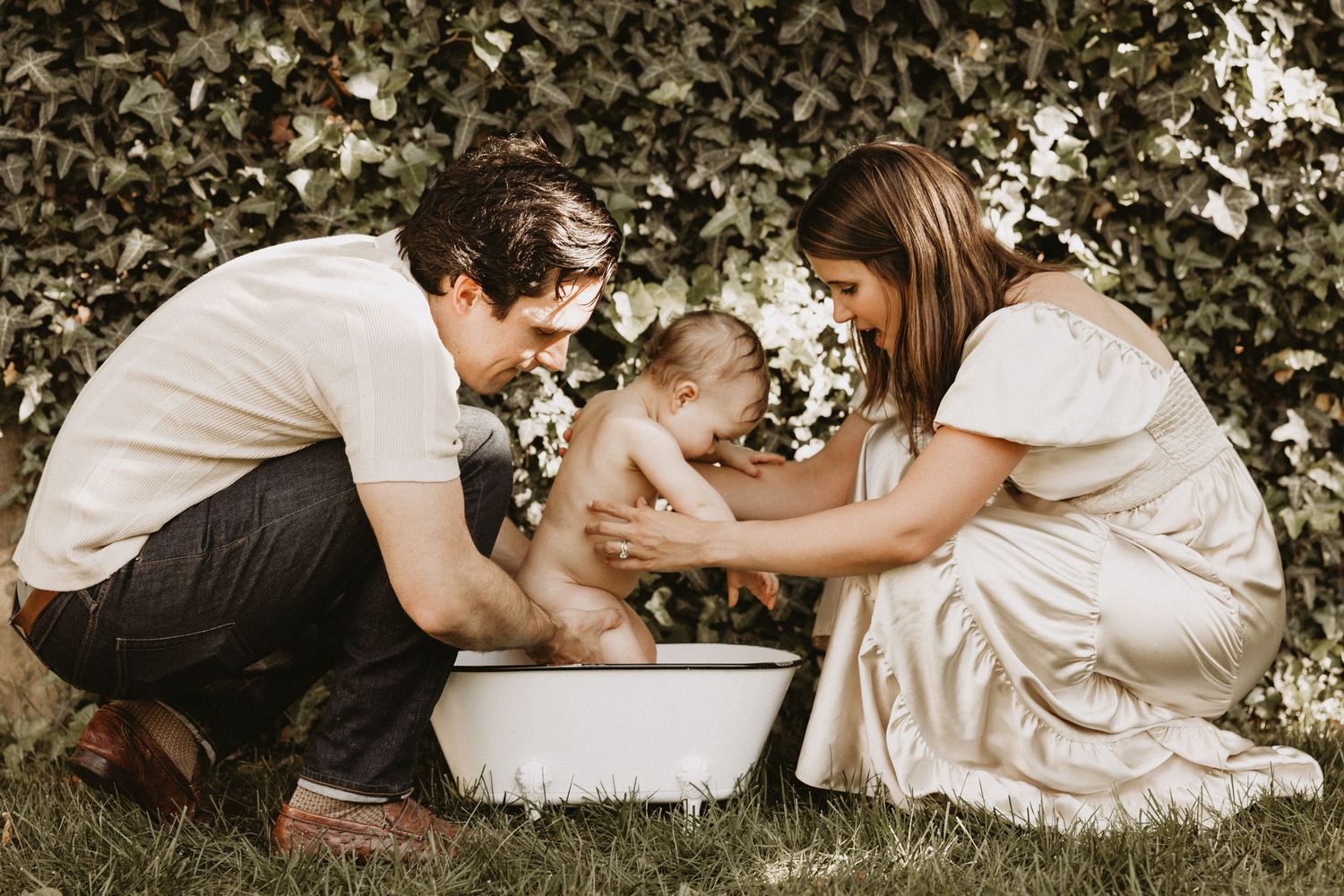 A couple in white clothing gives their puppy a bath in a basin outdoors with a green foliage background.