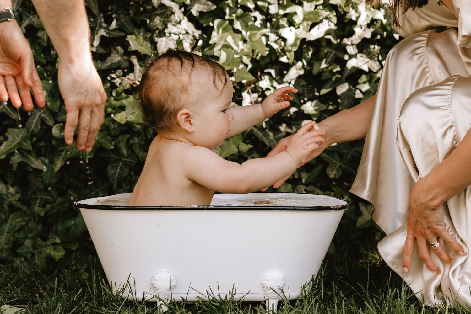 Two people enjoying a playful outdoor milk bath photo session with white porcelain tub in a natural garden setting.