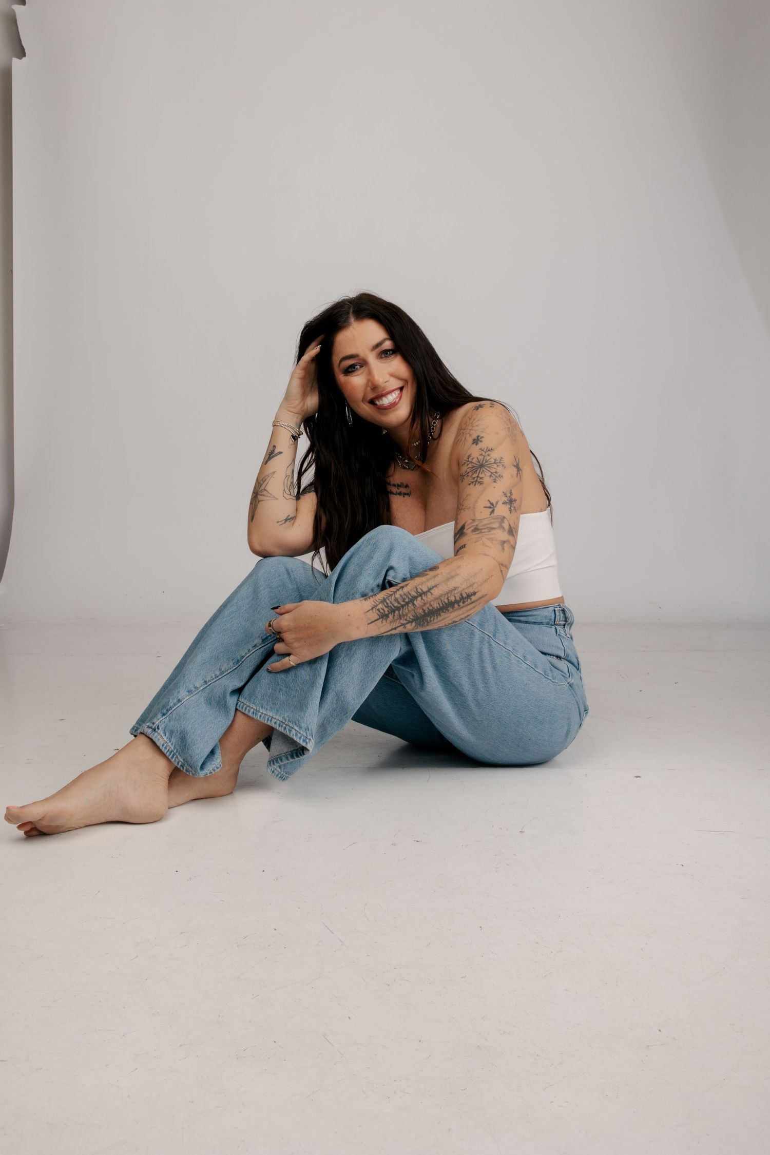 Casual portrait of person wearing blue jeans and showing tattoos while sitting barefoot on light gray studio background.