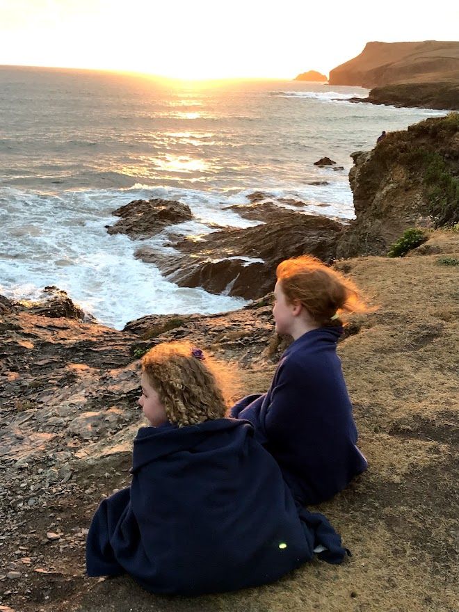 Two people sit at the edge of a rocky coastal cliff watching a dramatic sunset over the crashing ocean waves.
