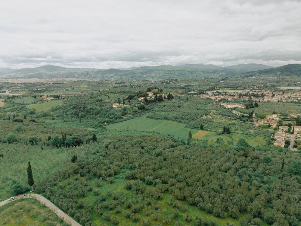 Aerial view of rolling Tuscan countryside with olive groves and scattered villas under cloudy skies.