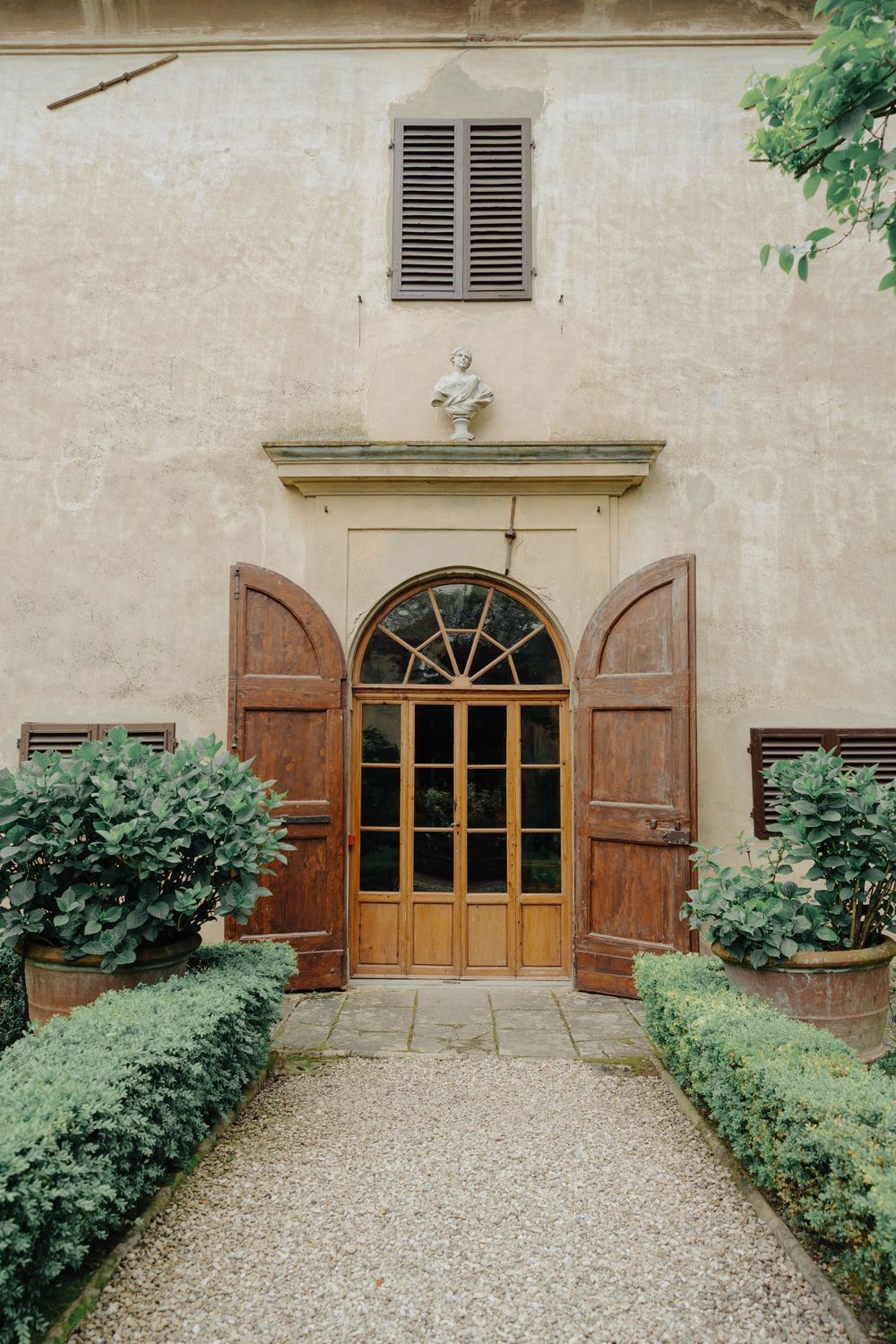 Elegant arched wooden doorway with antique shutters and potted plants along a gravel path at an Italian villa entrance.