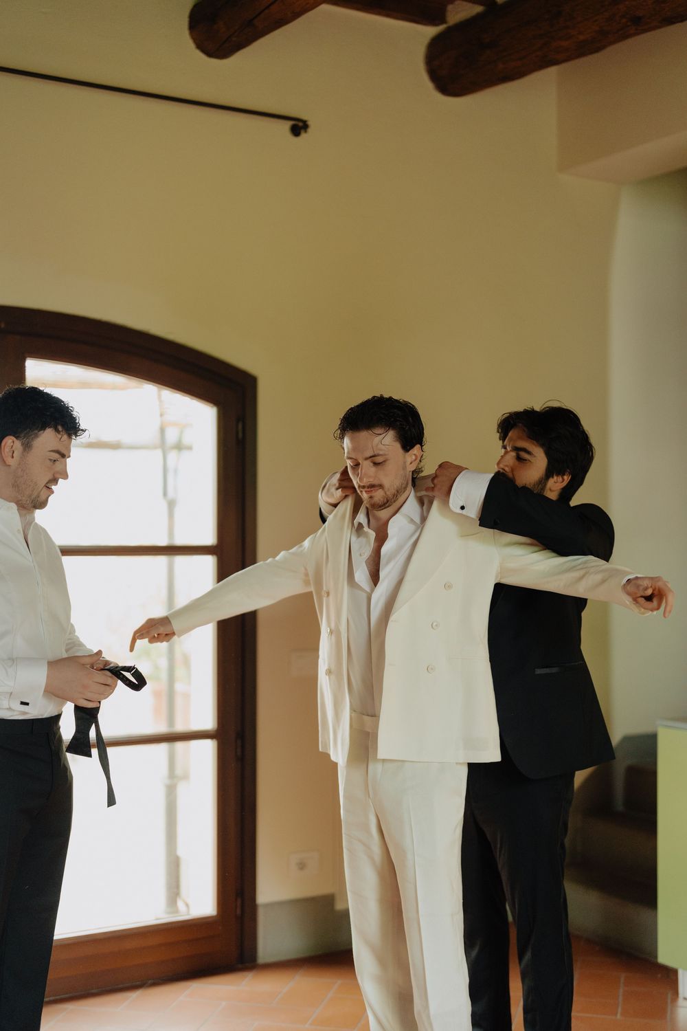 Groomsmen help the groom prepare by adjusting his white suit jacket in a rustic room.