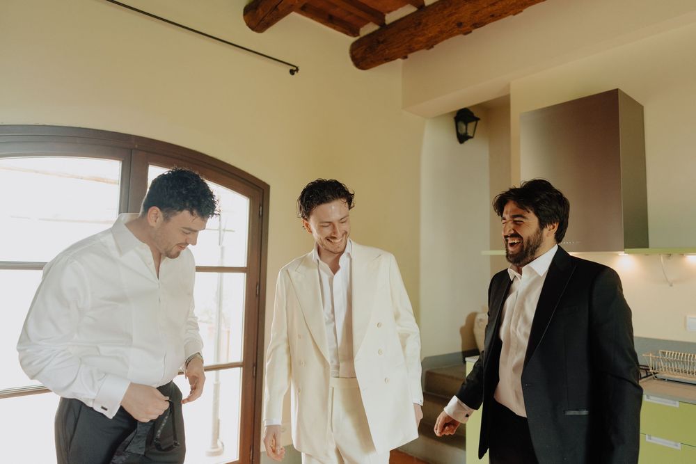 Three men in formal attire share a lighthearted moment while getting ready in a sunlit room.