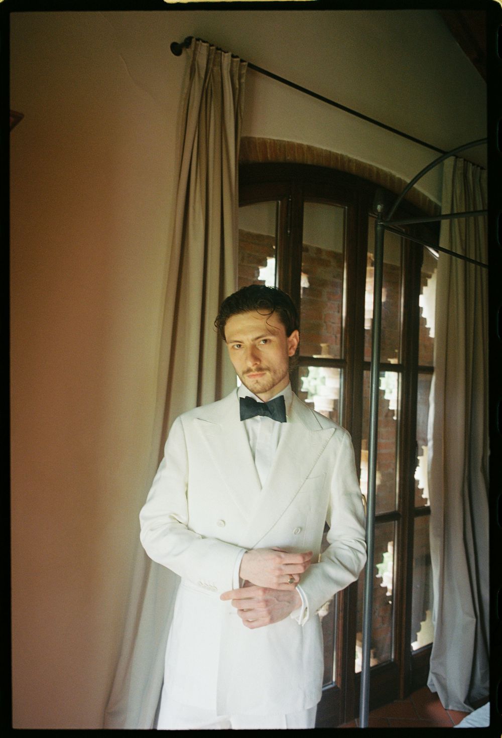 A person poses in a white tuxedo jacket and black bow tie against a backdrop with curtains and wooden furniture.