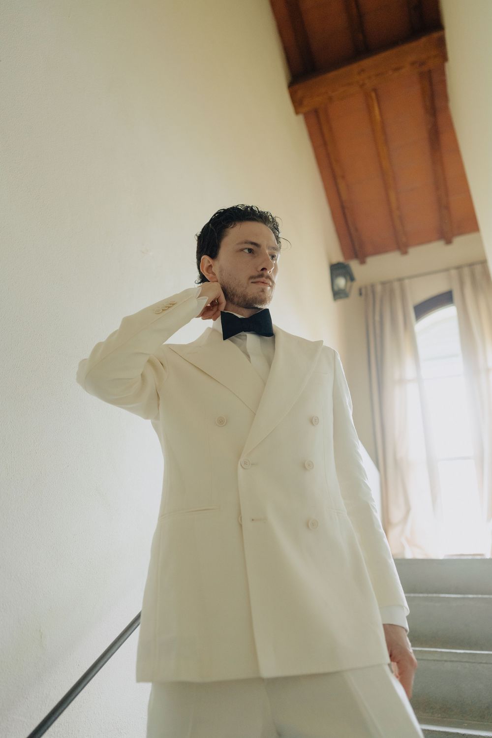 Elegant groom in a cream double-breasted tuxedo with black bow tie poses near a window in a classic interior setting.