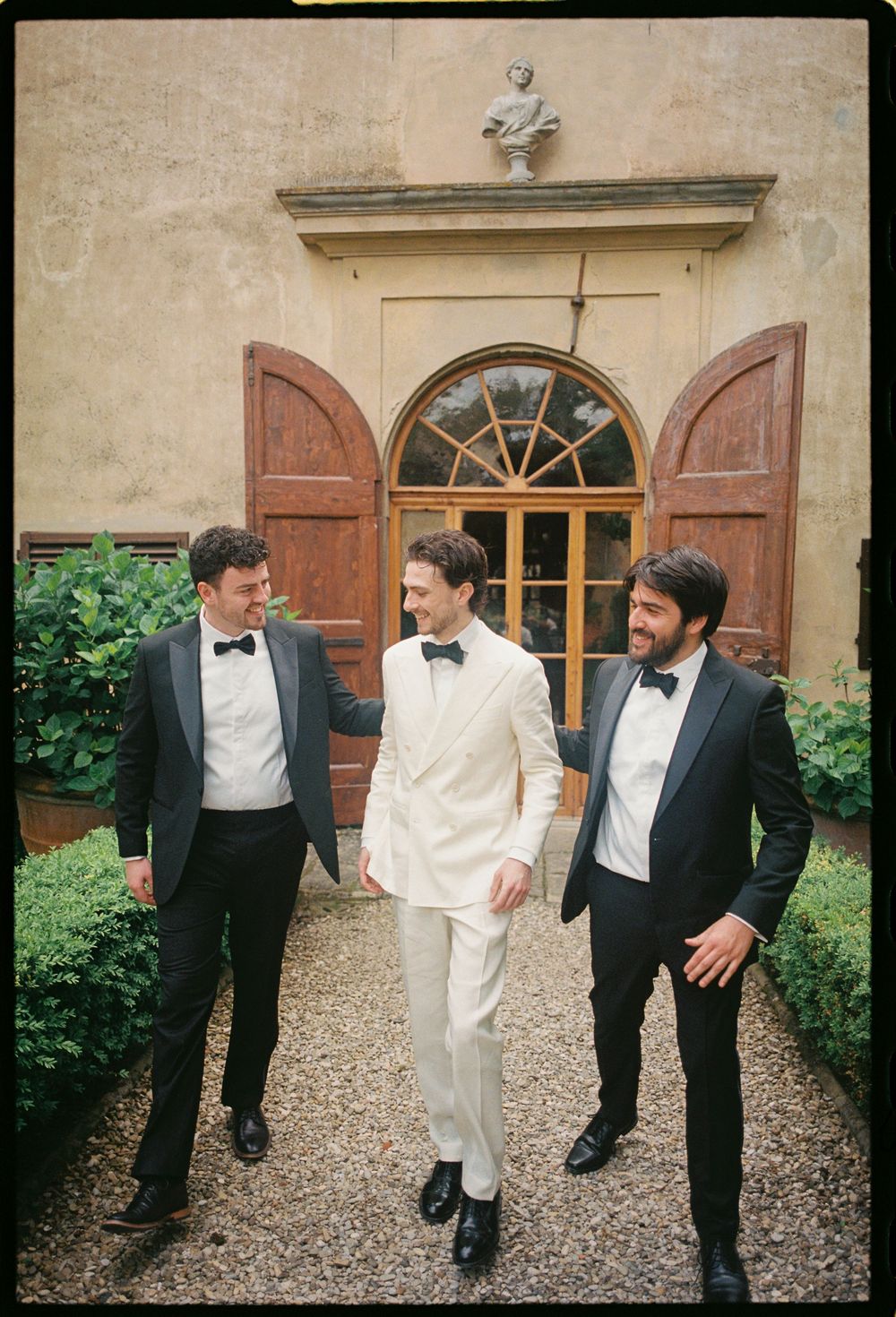 Three stylishly dressed friends in tuxedos sharing a laugh outside a historic Italian villa.
