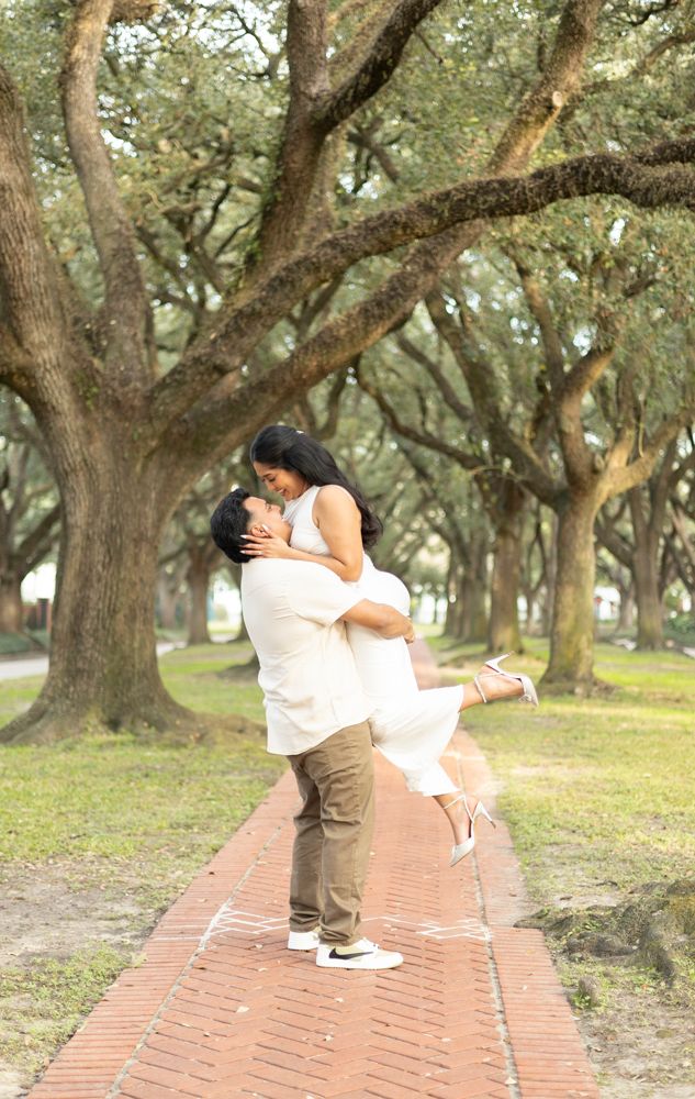 A couple embraces romantically on a brick pathway surrounded by mossy oak trees in a serene park setting.