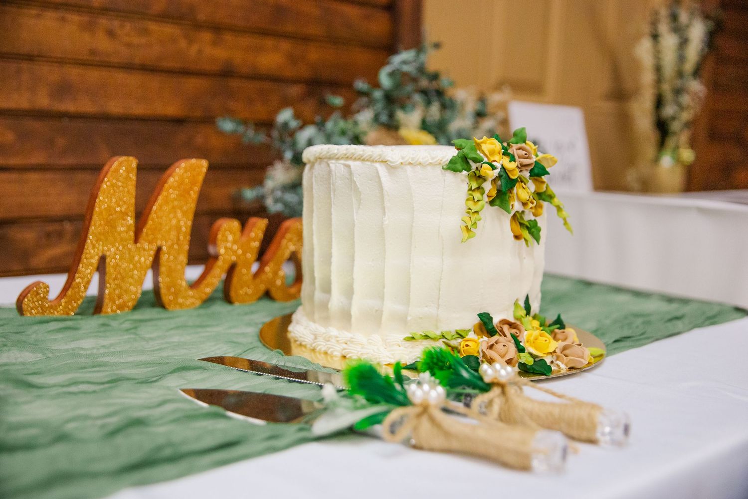 Small white wedding cake with yellow floral decorations sits on table with gold 'Mrs' sign and sage green tablecloth.