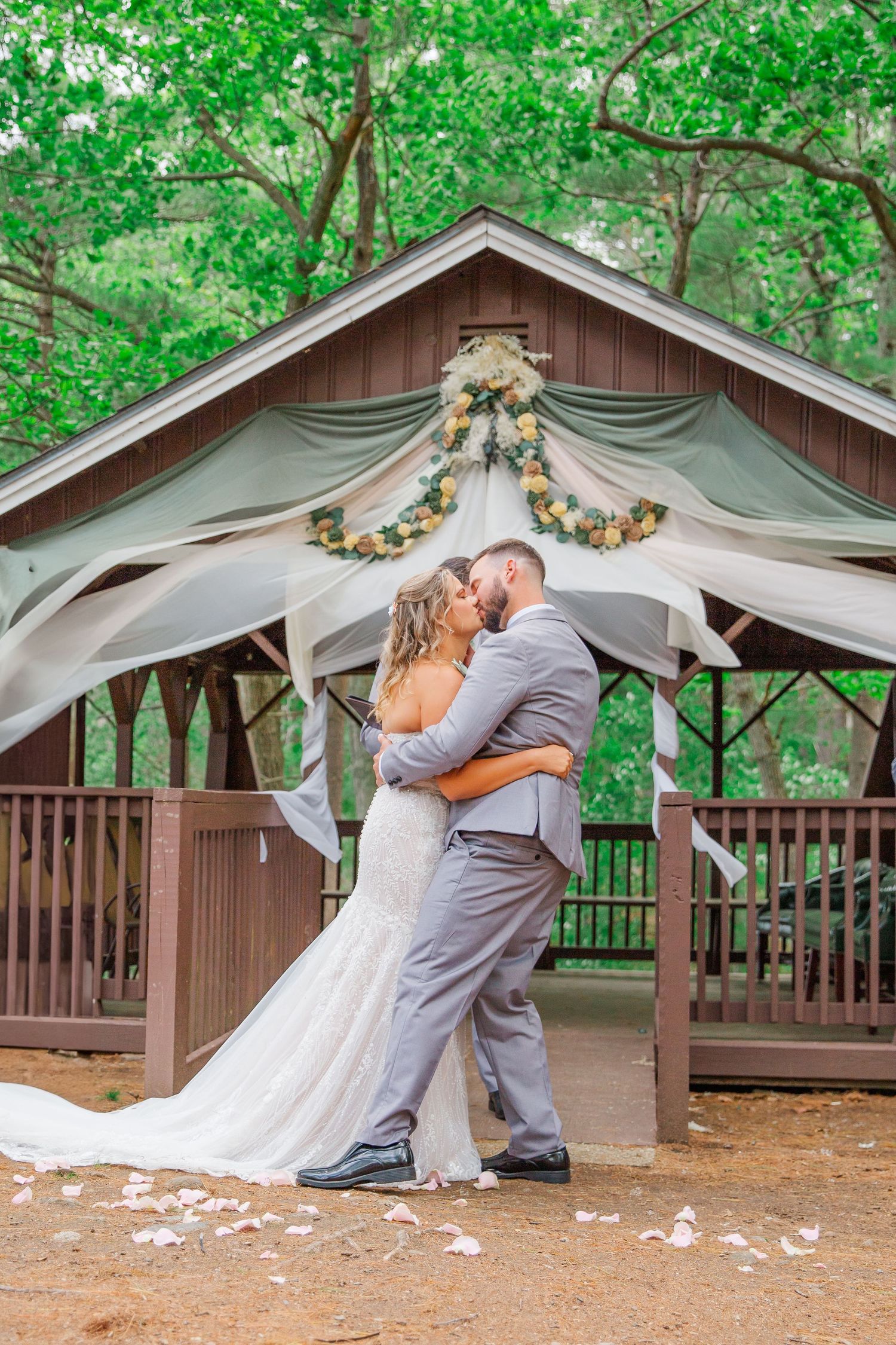 A couple embraces under a decorated wooden gazebo in a lush forest setting during their wedding ceremony.
