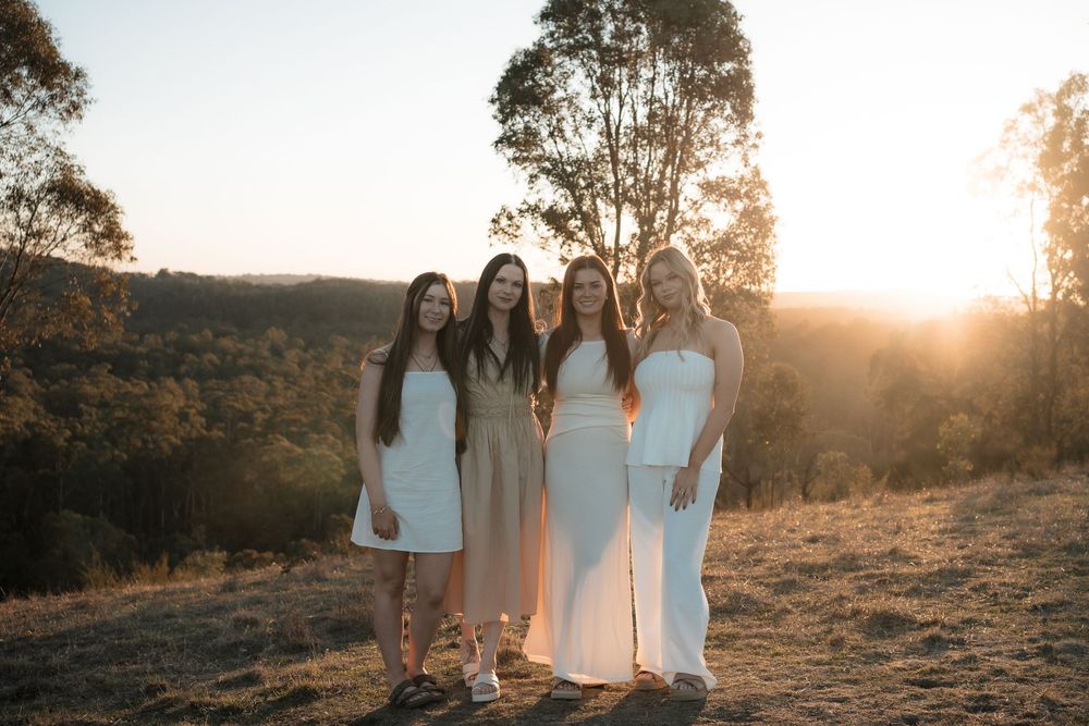 Four people in white dresses pose together at sunset in a natural outdoor setting with trees and rolling hills.