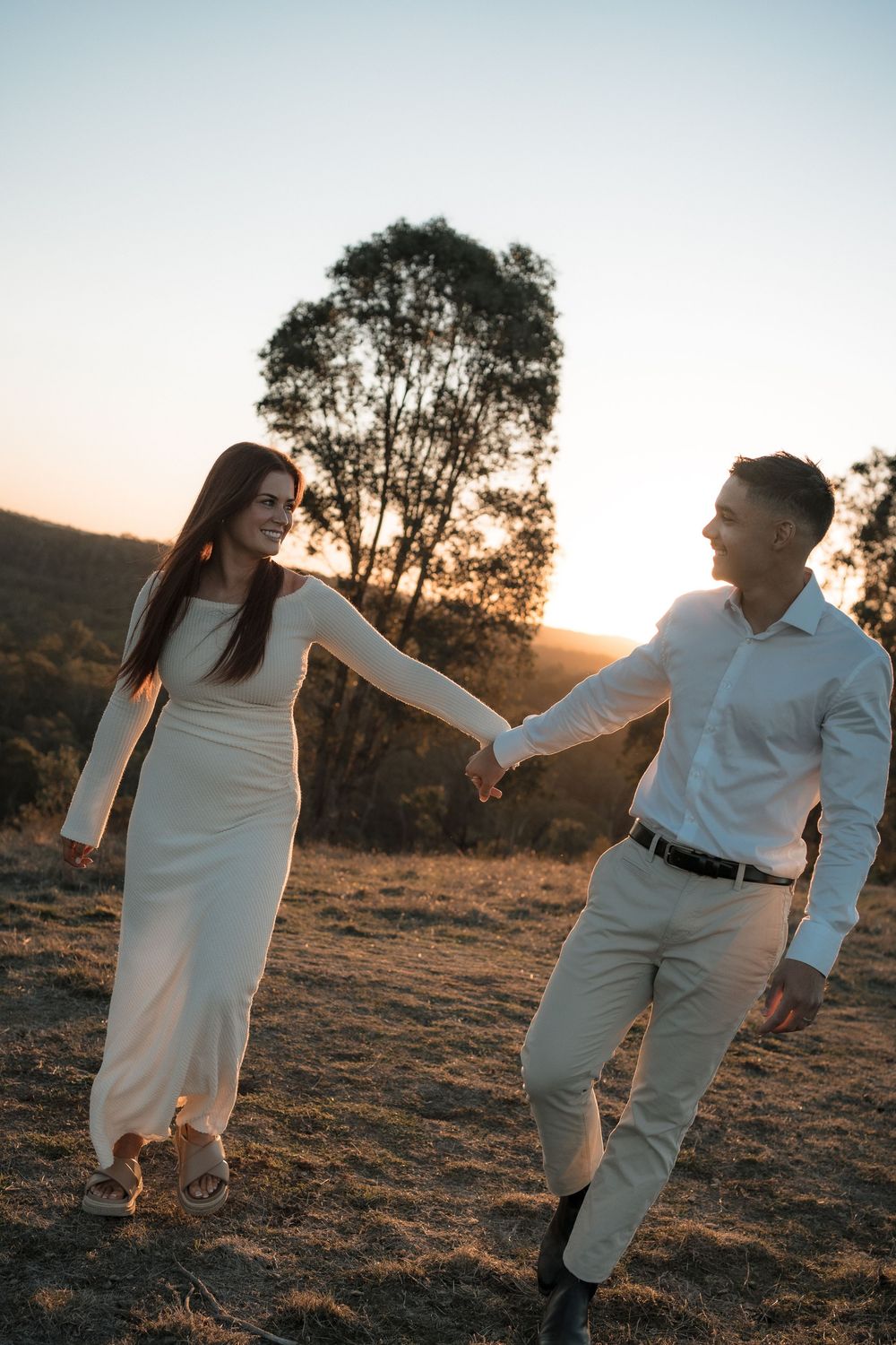 A couple in white outfits holds hands while walking along a hilltop at sunset with a tree silhouetted in the background.