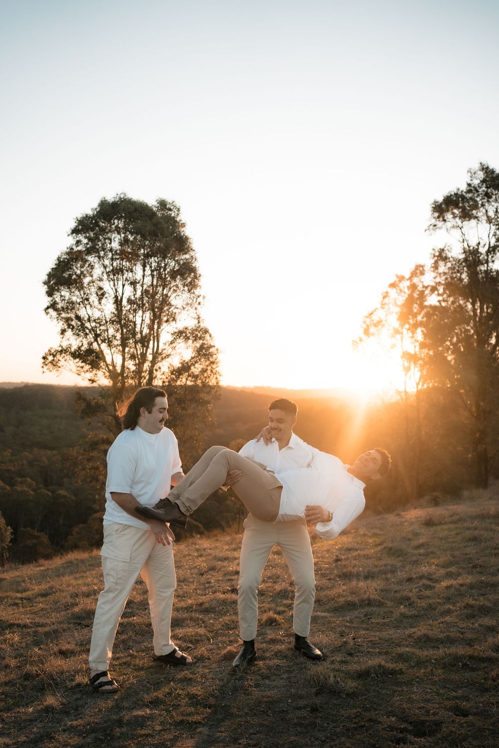 Two figures in white outfits stand together against a sunset on a grassy hillside with silhouetted trees.