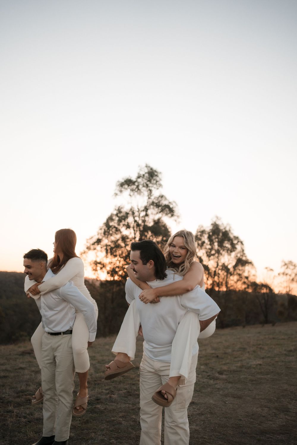 Two couples playfully giving piggyback rides at sunset in a natural outdoor setting with trees in the background.