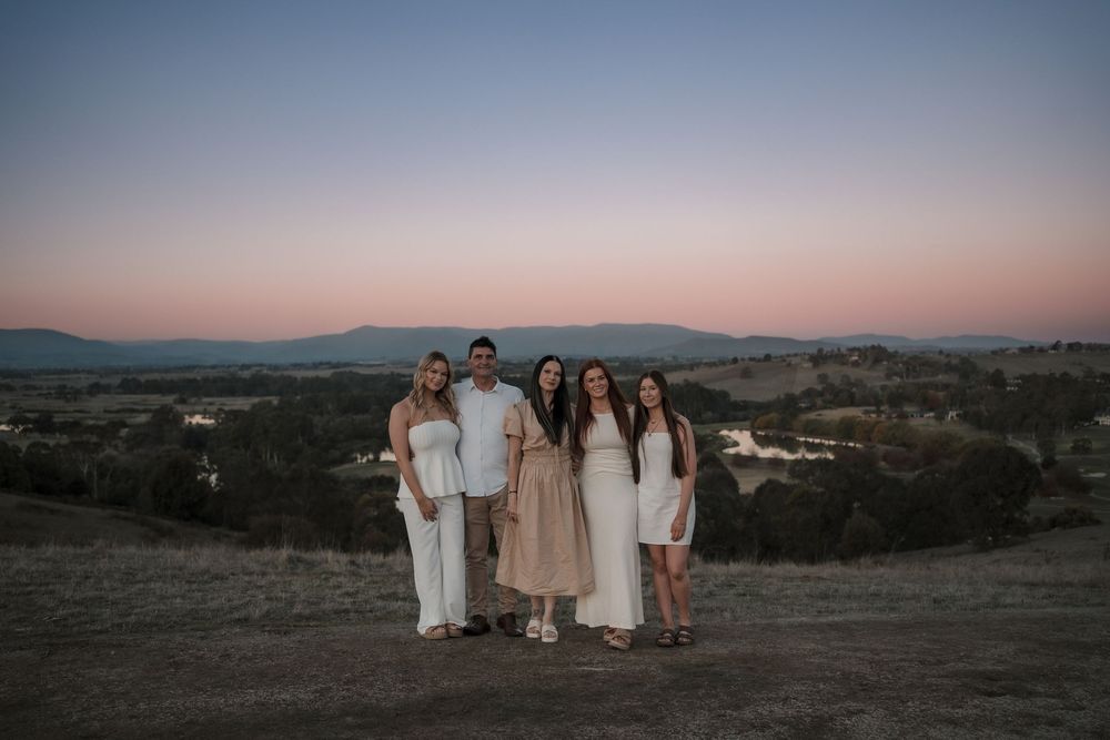 Group in coordinated white and beige outfits pose together on hilltop at sunset with mountain landscape in background.