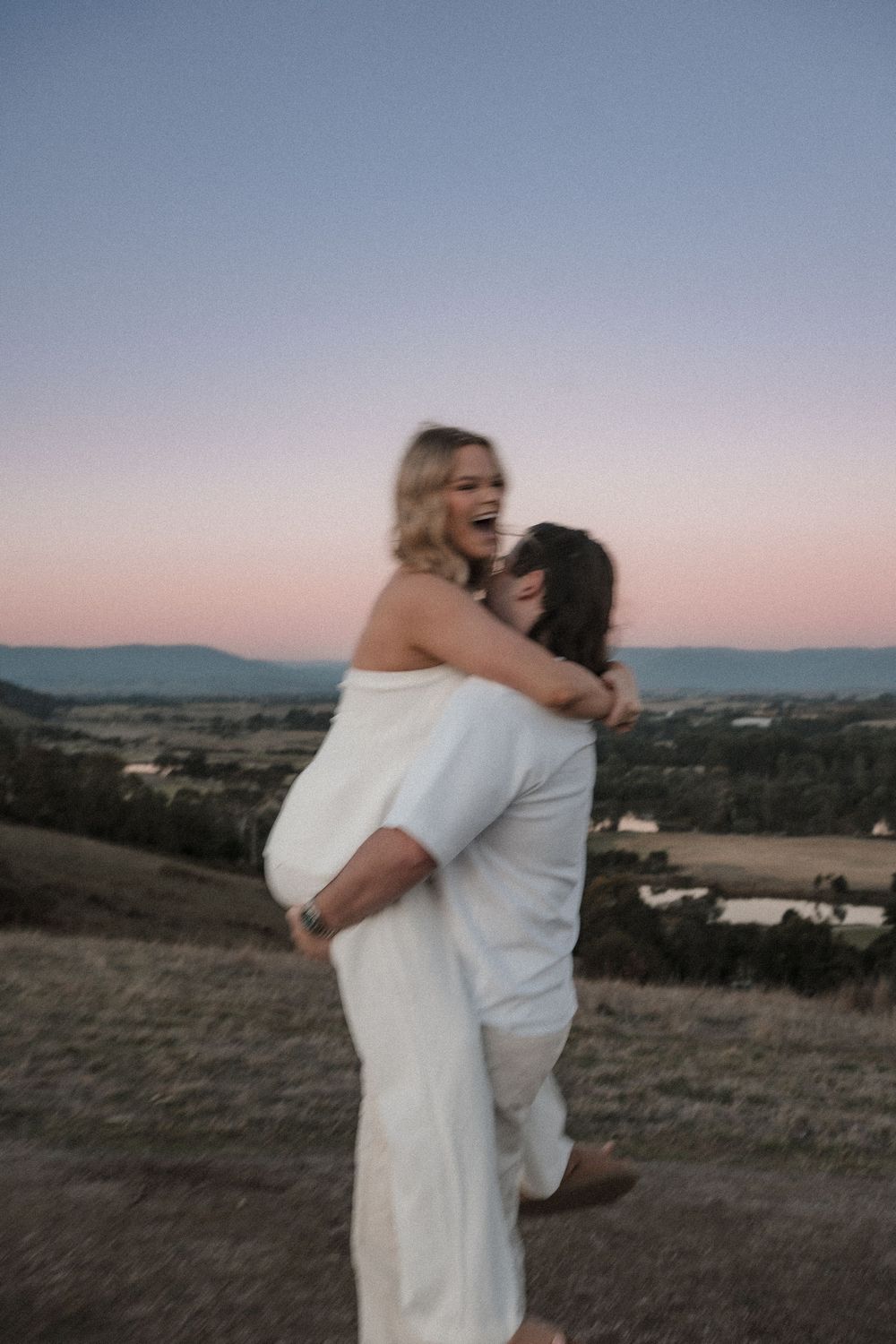 A couple shares a joyful embrace on a hilltop at sunset with rolling countryside and mountains in the distance.