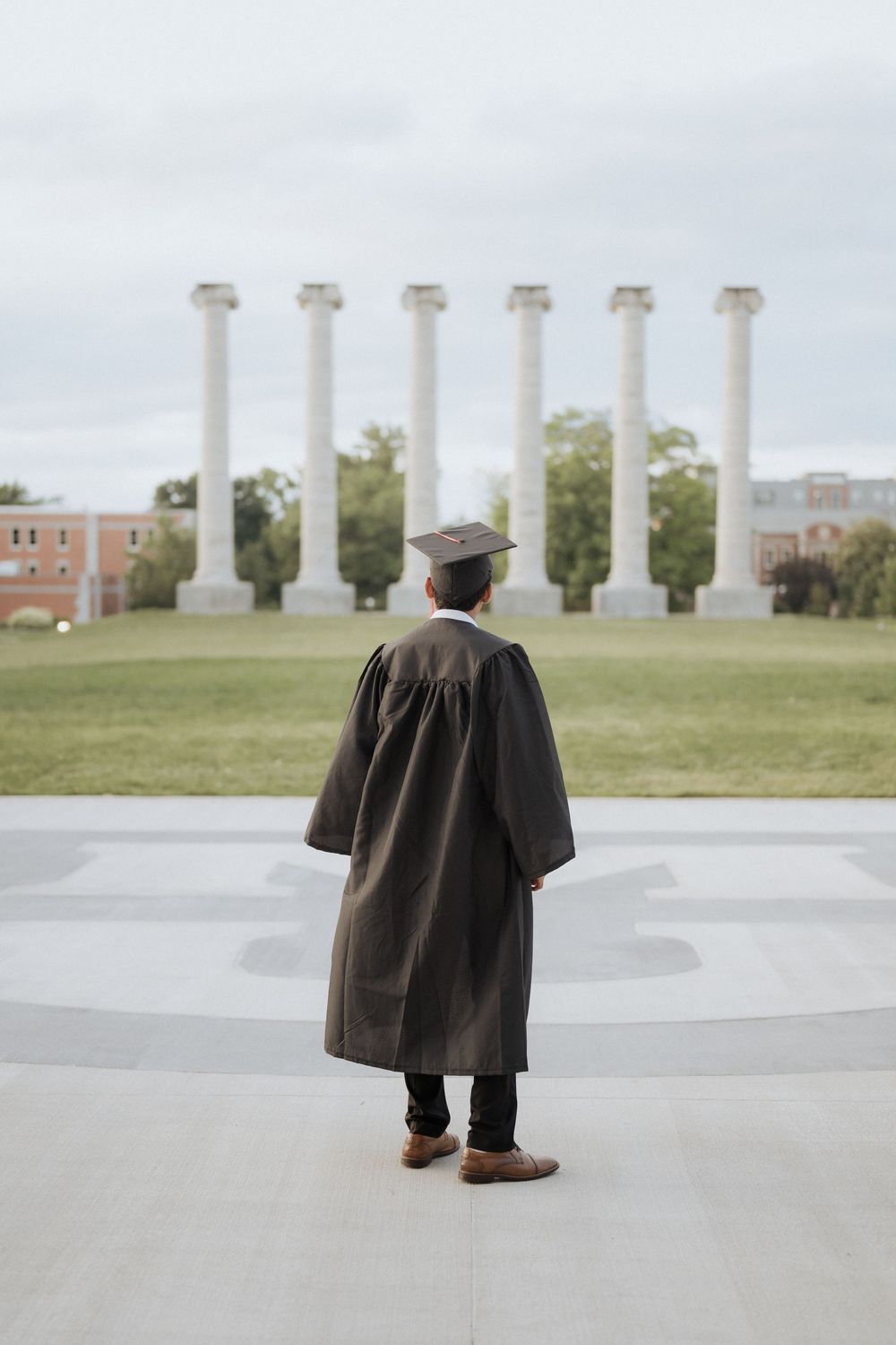 Graduate in black cap and gown walking past white columns on a university campus.