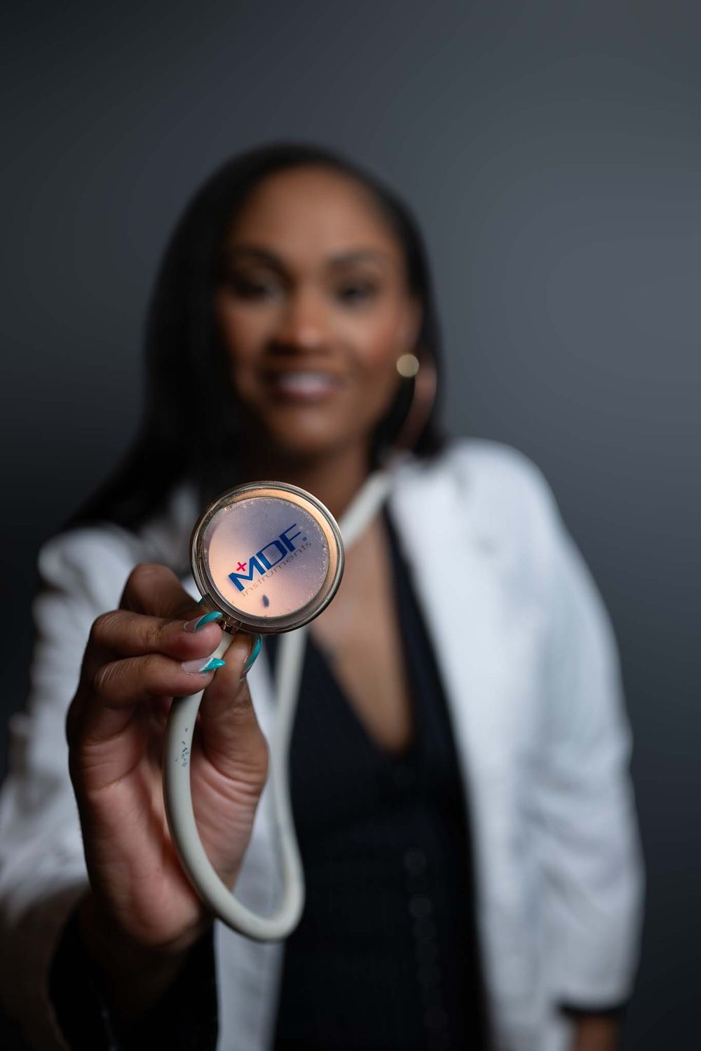 Healthcare professional in white coat holds up stethoscope with blue details against dark background.