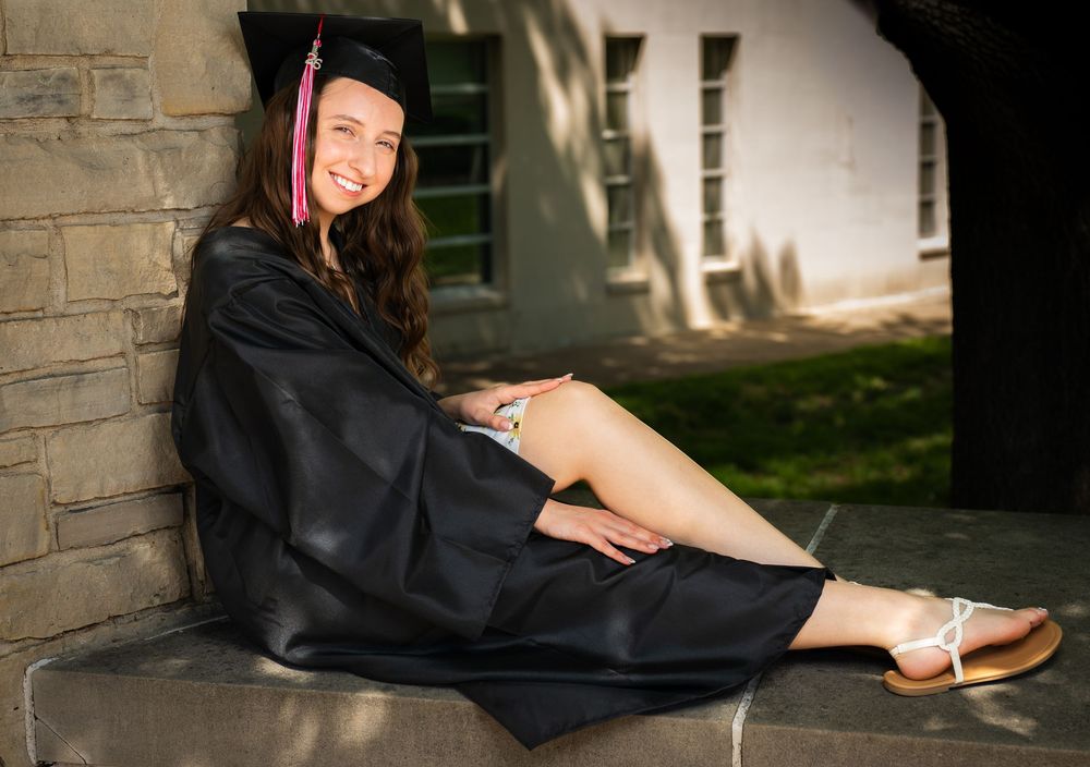 Smiling young woman in black graduation gown and cap sitting on stone steps outdoors on a sunny day.