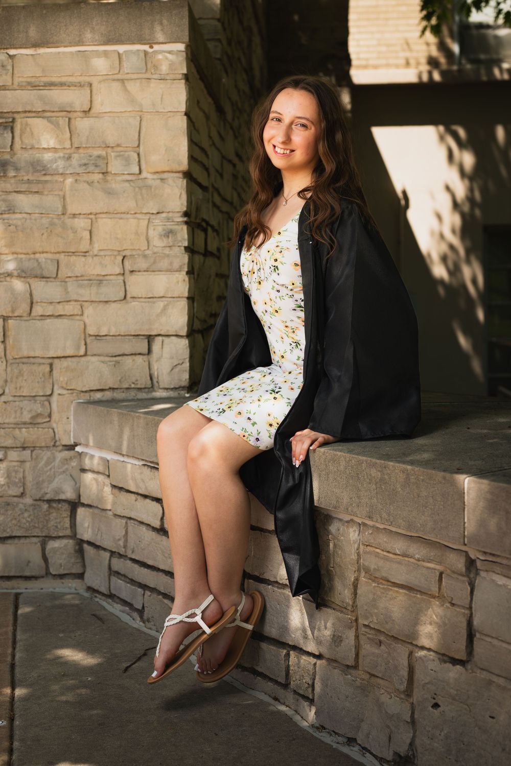 Young woman in floral dress and graduation gown sits smiling on stone wall outdoors in warm sunlight.