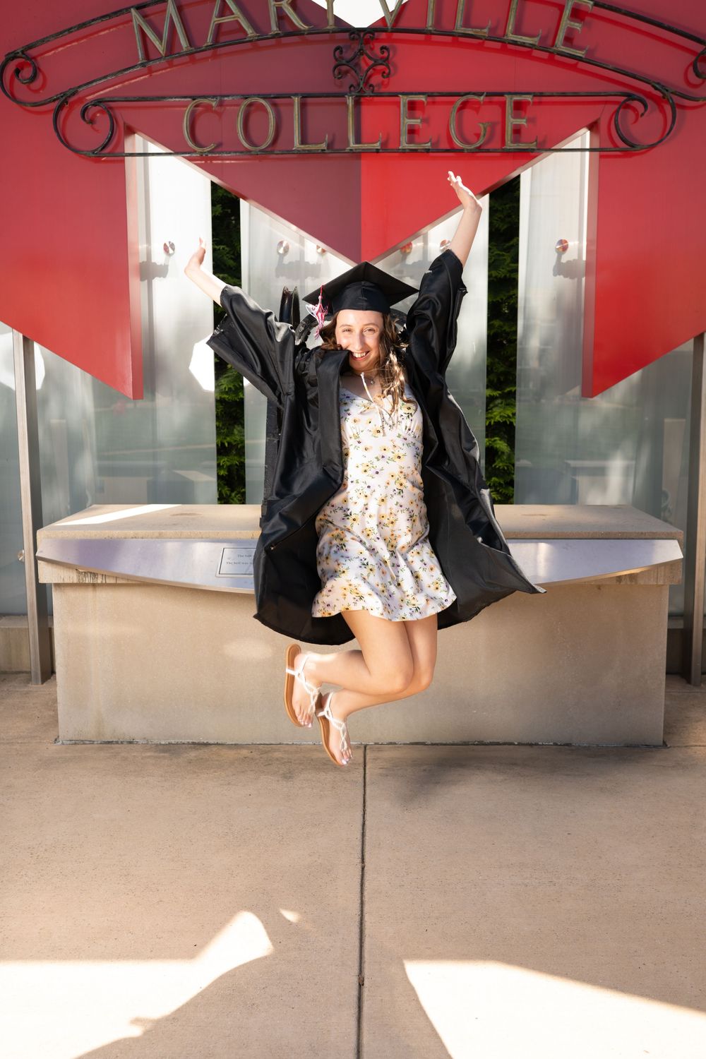 A joyful graduate jumps in the air wearing a cap and gown over a floral dress in front of Maryville College sign.