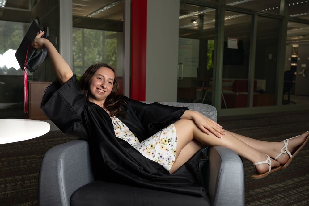 Young woman in graduation gown relaxing in chair, holding cap, smiling in modern building interior.