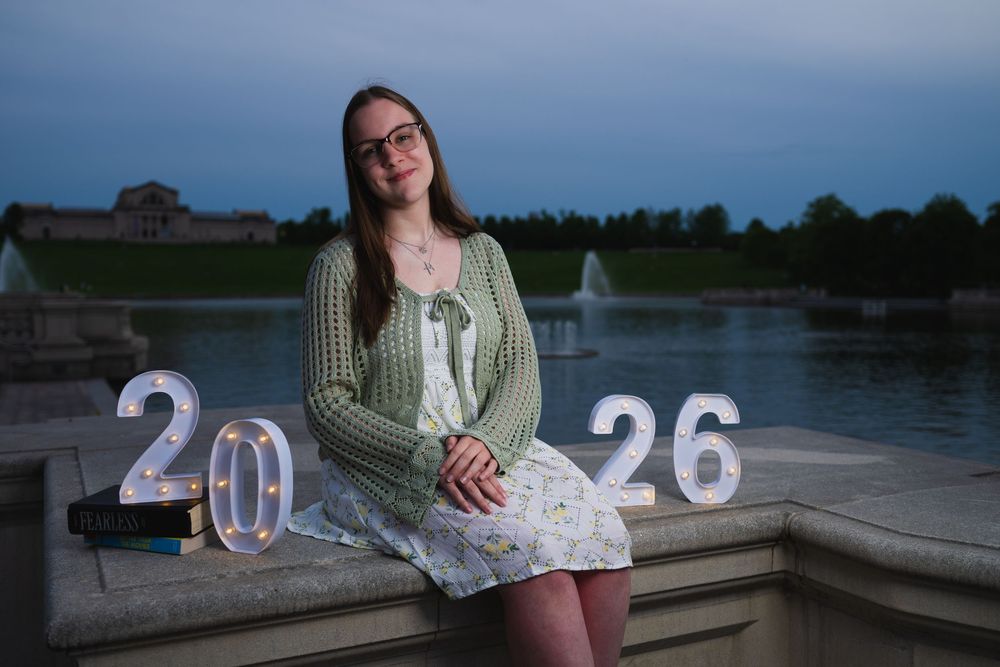 Young woman in green cardigan sits by fountain with illuminated 2026 signs at dusk.