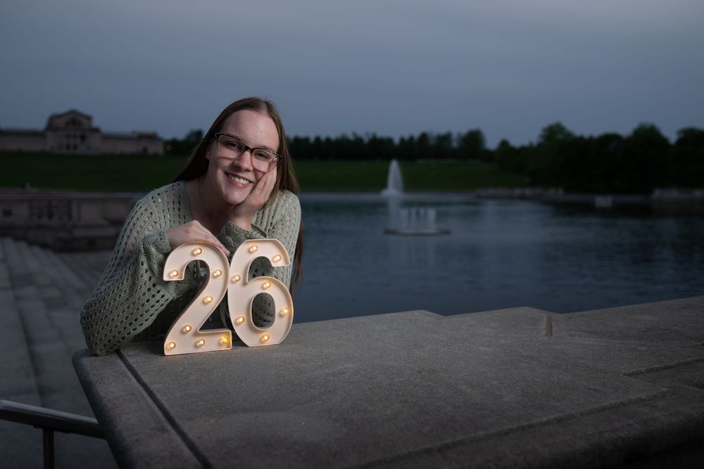 Woman smiling beside illuminated number 26 sign on ledge near fountain at dusk.
