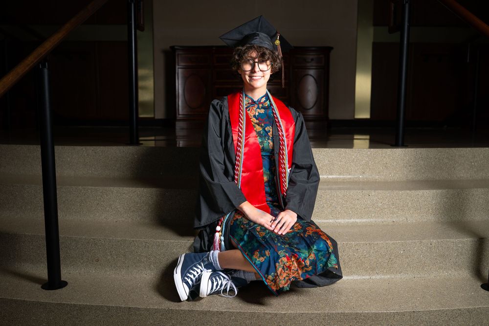 Graduate in cap and gown with red sash sits on stone steps wearing striped socks and sneakers, smiling at camera.